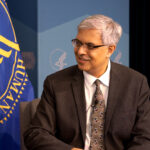 A man in a suit and tie sits and smiles, with a blue flag and a blue background featuring official emblems behind him.