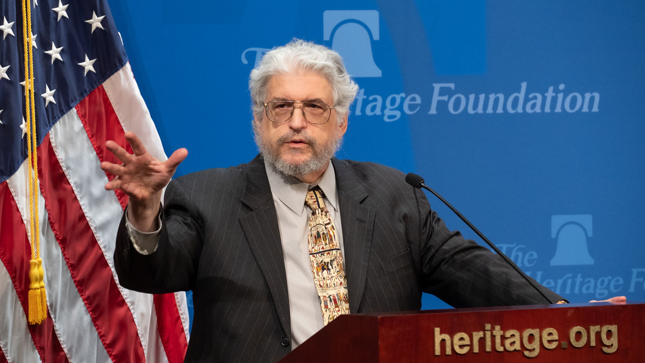An older man with gray hair and a beard speaks at a podium with a microphone, gesturing with one hand. An American flag and a Heritage Foundation logo are visible in the background.