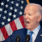 Joe Biden in a blue suit gestures angrily in front of an American flag while wearing an American flag pin.
