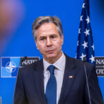 Antony Blinken in a blue suit purses his lips in front of an American flag and a NATO symbol.