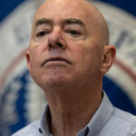 Homeland Security Secretary Alejandro Mayorkas look ahead in a light blue button down shirt during a press conference with the DHS flag behind him.