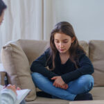 Preteen girl in jeans and a sweater crosses her hands on a couch as a therapist asks her questions with a pad and paper.