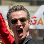 Gavin Newsom emotes with mouth open. People hold signs in rainbow font saying "Gavin."