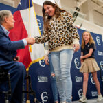 Governor Greg Abbott wears a navy blue suit, sitting in his wheelchair, while he shakes the hand of a teenaged girl wearing a cheetah print sweater.