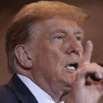 Former President Donald Trump in a suit at a microphone with his hand raised, Finger pointing upward