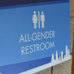 A dark and light blue bathroom sign says "ALL-GENDER RESTROOM" with two white man and woman images, side-by-side.