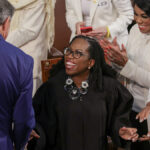 Ketanji Brown Jackson shakes hands with a man in a blue suit while she wears a large necklace above her black robes