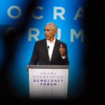 Former President Barack Obama stands at a podium and speaks in a black suit and white shirt without a tie.