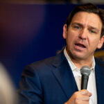 Ron DeSantis in a blue suit holds a microphone