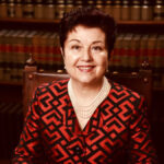 Virginia Prodan sits in front of bookshelves wearing a blouse and pearl necklace.
