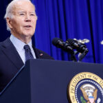 President Biden speaks at a podium in a dark suit with a royal blue curtain behind him.