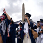 Students wear graduation caps and masks in front of the Washington Monument.