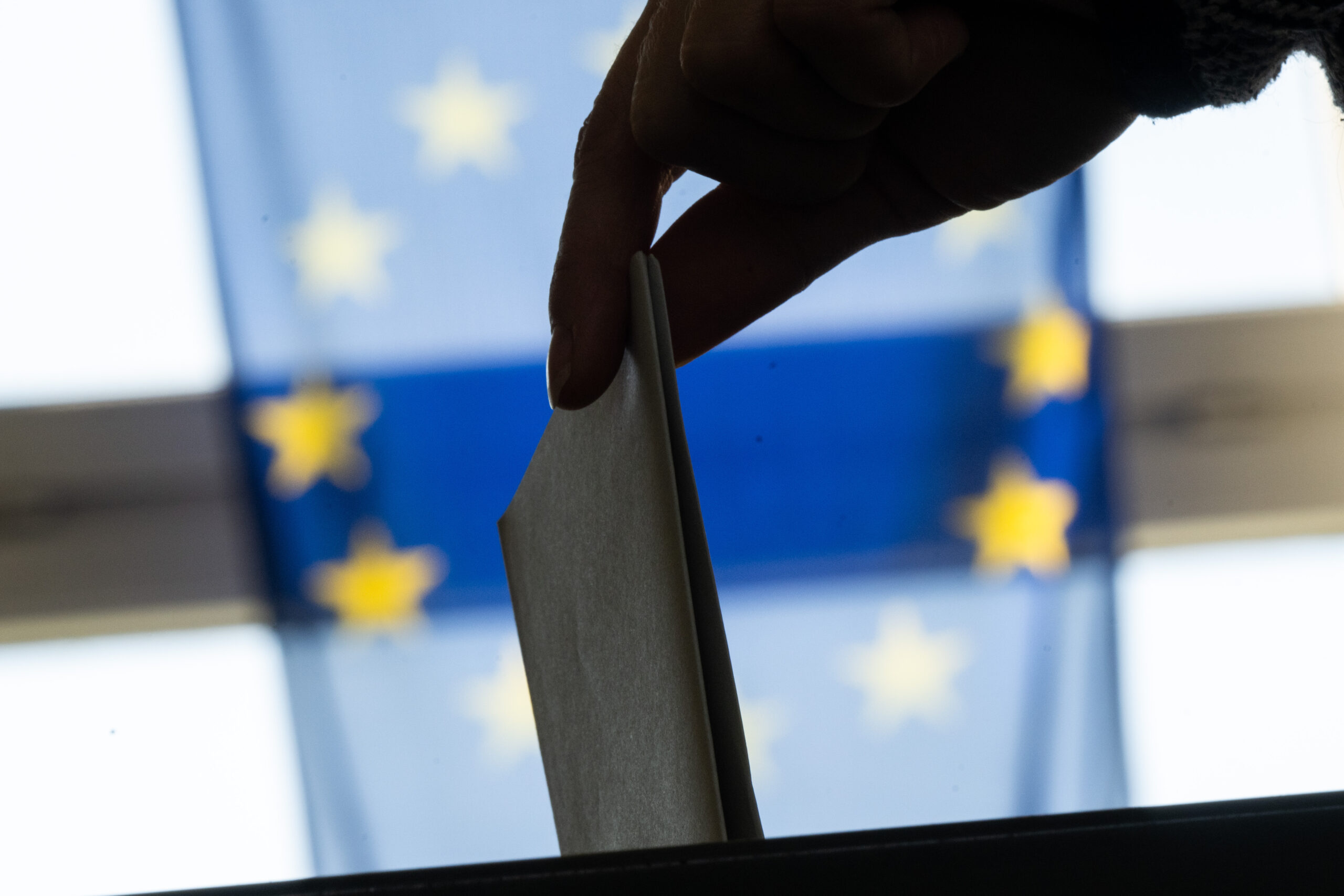 A woman's hand drops a paper ballot into a ballot box in front of the European Union flag.