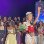 Trans-identifying male contestant waves waves from the stage in a gown, crown, and flowers with other contestants in the background