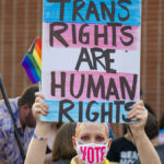 Woman holds sign reading "Trans Rights Are Human Rights" while wearing a face mask with the text "Vote."