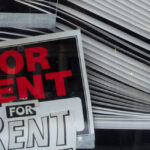 A crooked "For Rent" sign in the corner of a window with dilapidated blinds.