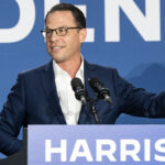 Josh Shapiro in a blue suit raises his hind behind a podium reading "Harris"