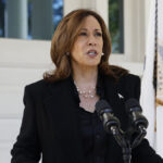 Kamala Harris in a black suit stands behind a microphone in front of an American flag.