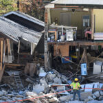 Destroyed homes sit on a rocky riverbed, a worker in neon helping with the clean-up effort.