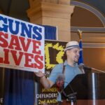 A man wearing a hat holds a sign that states, "Guns Save Lives."