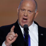 Tom Homan in a dark suit and tie speaking on stage forcefully with his finger pointing forward