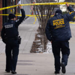two police officers in Metropolitan Police Department jackets walk down a sidewalk with their backs to the camera holding up police caution tape as they walk under it to a crime scene