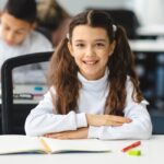 A young girl with a ponytail sits at a desk with her hands folded in front of her.