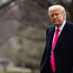 Donald Trump in a blue suit with a red tie walks outside