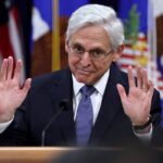 Merrick Garland stands at a podium and puts both his hands up in the halt position during a press conference.