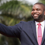 Byron Donalds wears a dark grey suit and red necktie as he smiles.