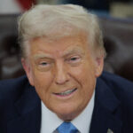 Donald Trump smiles, wearing a dark blue suit and light blue necktie.