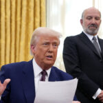 Commerce Secretary Howard Lutnick looks on as President Donald Trump sits behind the Resolute Desk in the Oval Office.