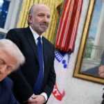 Howard Lutnick stands with his hands folder as Donald Trump signs an executive order.