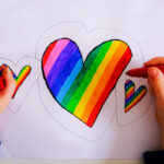 a child's hands holding a marker coloring in hearts with the LGBTQ rainbow