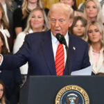 Donald Trump in a blue suit and red tie at the podium gesturing toward the crowd with several women standing behind him