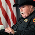 Sheriff Bill Waybourn wears a black cowboy hat and uniform while sitting in front of an American flag.