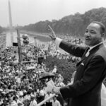 in a black-and-white photo, Martin Luther King Jr. stares across a crowded Washington Mall before a speech with photographers taking pictures of him in the background