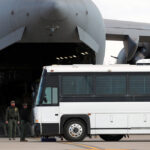 Large white bus parked next to the back of large cargo plane, its cargo door open wide.