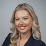 A young woman with long blonde hair and blue eyes, wearing a dark blazer and a gold cross necklace, smiles at the camera against a plain light gray background.