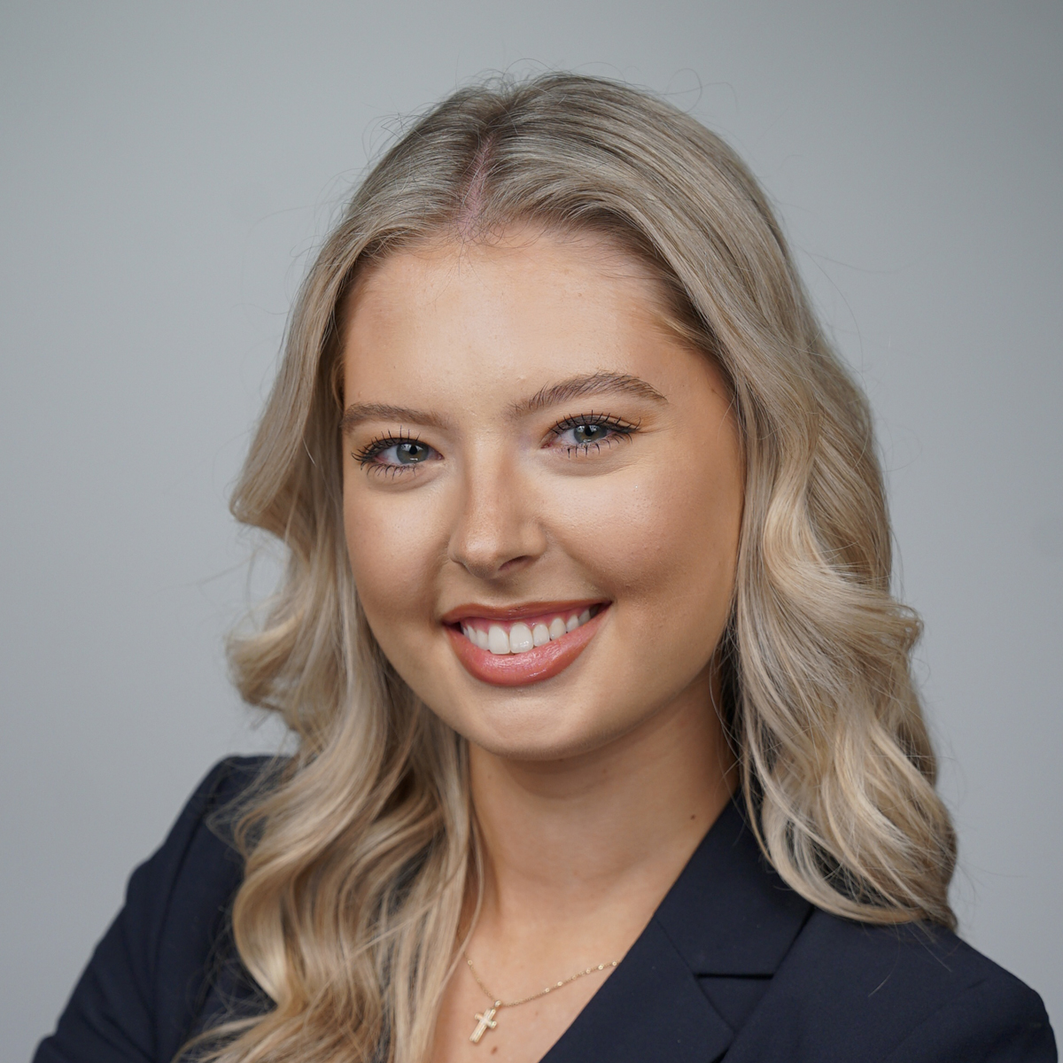 A young woman with long blonde hair and blue eyes, wearing a dark blazer and a gold cross necklace, smiles at the camera against a plain light gray background.