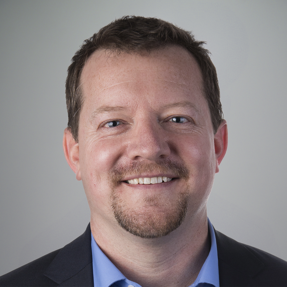 Smiling man with short brown hair, light beard, and mustache, wearing a dark blazer and light blue collared shirt, posed in front of a plain light gray background.