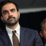Zohran Mamdani in a blue suit and brown tie stands in front of a dark background, looking serious, Two other people, a woman and a man, are partially visible behind him.