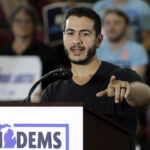 Michigan Democrat Abdul El-Sayed compared circumcision to transgender surgery. Pictured: El-Sayed, a man speaks at a podium labeled MIDEMS in front of an audience. He gestures with one hand while addressing the crowd. People in the background hold signs and appear to be listening attentively.
