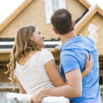 Homeownership concept. Pictured: a couple stands in front of a partially built house, embracing. The man points toward the home while the woman holds rolled blueprints, both looking at the construction with anticipation.