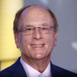 BlackRock is among the companies accused of merely rebranding DEI efforts or preserving them altogether. Pictured: CEO Larry Fink, a smiling middle-aged man with glasses and thinning hair, wearing a navy blue suit and light-colored tie, stands in front of a blurred background with hints of yellow, blue, and a partial white star.
