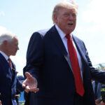 Donald Trump in a suit gestures with open arms outside, with the U.S. Capitol building in the background and people standing nearby.