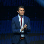 Charlie Kirk, dressed in a blue suit and red tie, stands on stage, clapping his hands, with a dark blue background and a spotlight shining from the upper right corner.