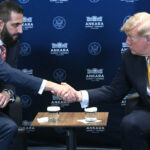 Donald Trump and Syria's president in suits sit across from each other, shaking hands over a small table with coffee cups. Flags and a blue backdrop with ANKARA and emblems are behind them.