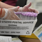 A person wearing gloves handles a USPS tray filled with mail-in ballots. The tray is labeled HALF MM TRAY – EIRS 74HP. Other paperwork is visible in the foreground.