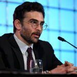 Hasan Piker in a suit and glasses speaks into a microphone at a panel, with his hands clasped on the table. Blue and orange backgrounds are behind him.